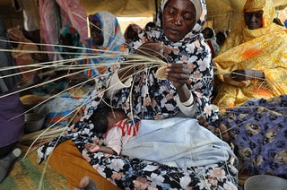 A woman with a child in her lap weaves a basket at the Abu Shouk Women's Center in Sudan, Africa.