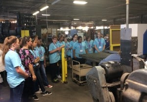 A group of young women in matching t-shirts visit the floor of a manufacturing plant