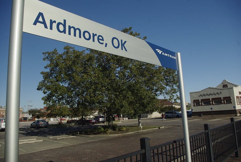 A narrow metal sign says, "Ardmore, OK, Amtrak". The Ardmore Oklahoma railroad depot is in the background