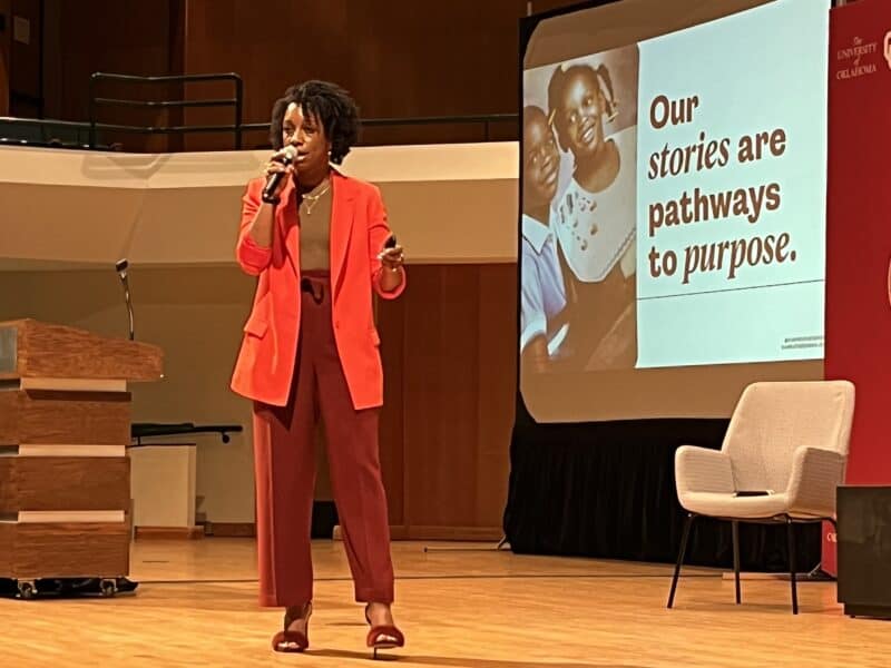 A woman speaking on stage. She has dark skin and dark hair in a natural style. The slide on screen shows her as a child with her brother, and the text says, "Our stories are pathways to purpose." 