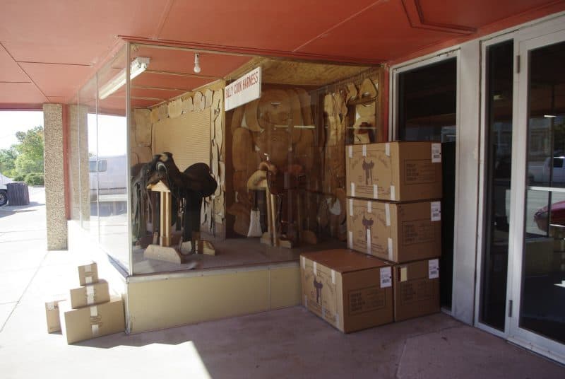 A converted retail building in the center is used by a saddle maker for storage and warehousing.  A window display shows saddles and the molds used to make them.  A sign says "Billy Cook armor." Several boxes of saddles and accessories are outside ready to be picked up by the delivery company. 
