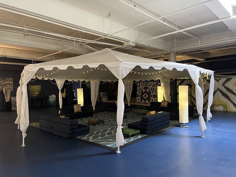 A large room, with a pavilion tent draped with string lights. Comfortable lounge chairs arranged on a rug. In the background, the walls are hung with fabric art in earth tones