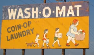 A faded sign says, "Wash-o-mat coin-op laundry". Painted figures of a family are carrying their basket, soap and bleach and are followed by a frisky dog.