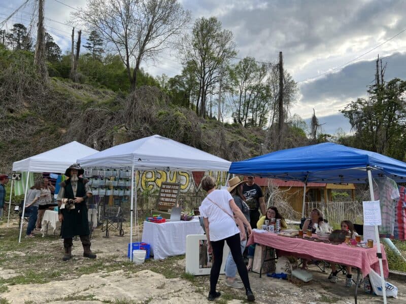 On a hillside lot overgrown with vines and weeds, a flat place has been cleared and three artists' booths are set up with their creative works for sale. Customers and one person dressed in roguish garb are visiting the booths. 