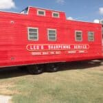 A travel trailer painted red and decorated as a railroad caboose has lettering that says, "Leo's sharpening Service"