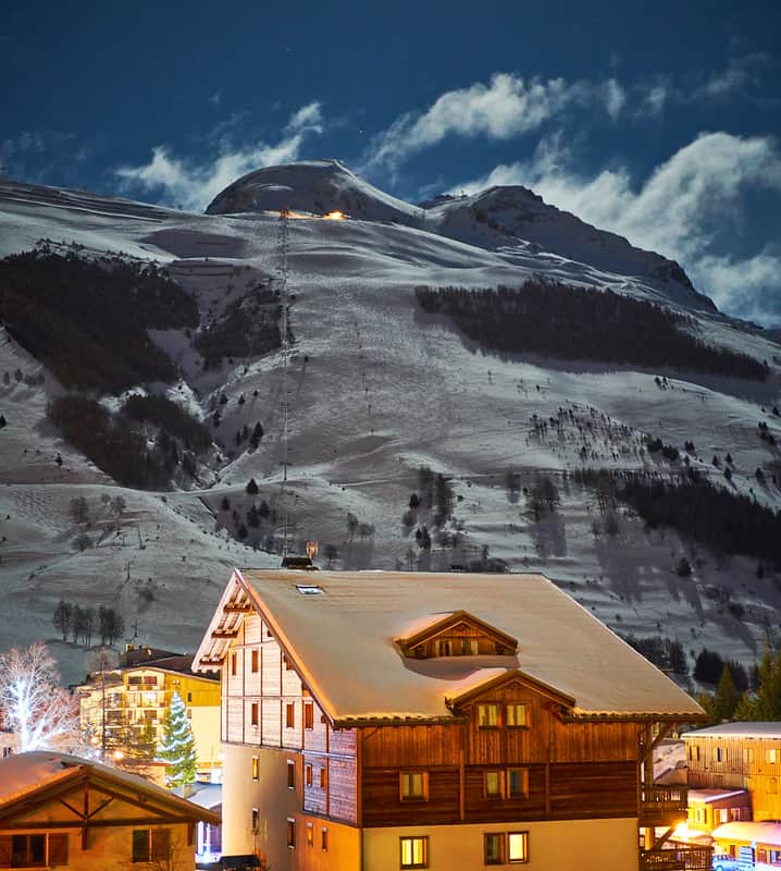 The moon rises over a dimly lit snowy mountain, as the ski resort below is warmed with the glow of street lights and interior lights. 