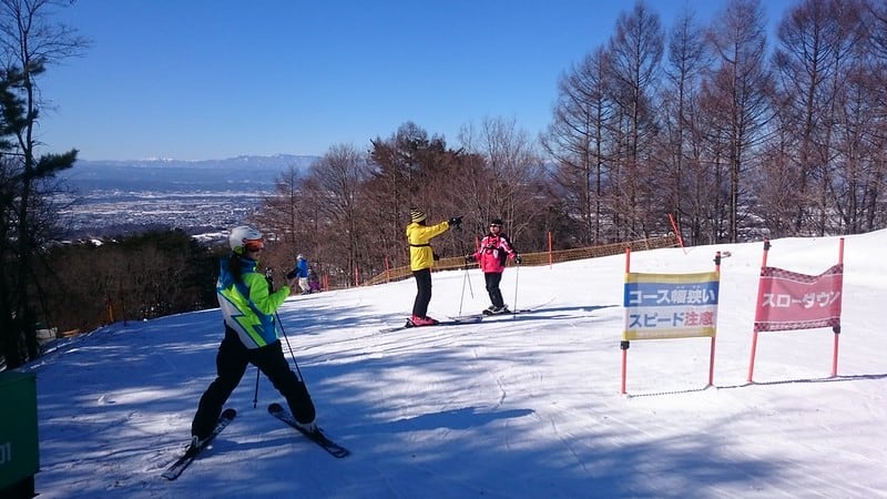 A group of skiers in colorful outfits on snowy slopes. Banners in Japanese script are in the foreground.