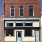 A two-story brick downtown building has been recently rehabbed with new windows and updated paint. A small "for sale" sign is in the window.