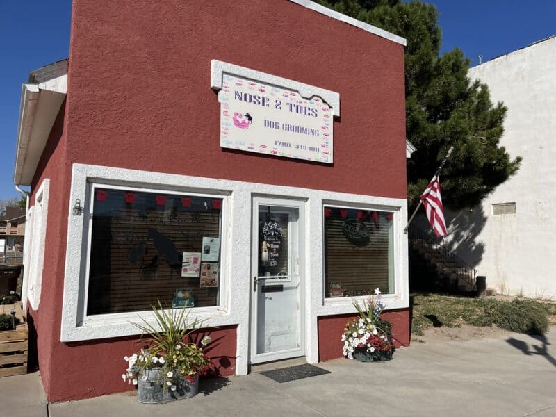 A red storefront building in Stratton, Colorado, is filled with a dog grooming businesses. Flower pots out front and a flag on the building add a friendly vibe. 