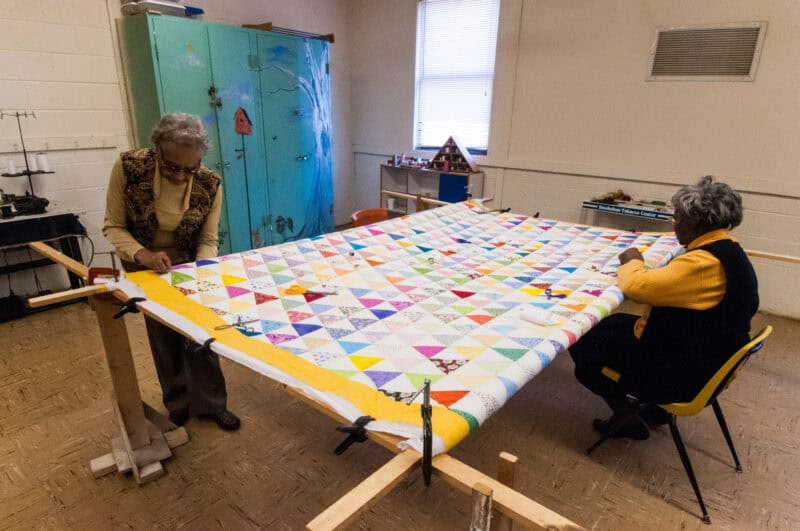Two women with dark skin and white hair are working on a quilt in a quilting frame. 