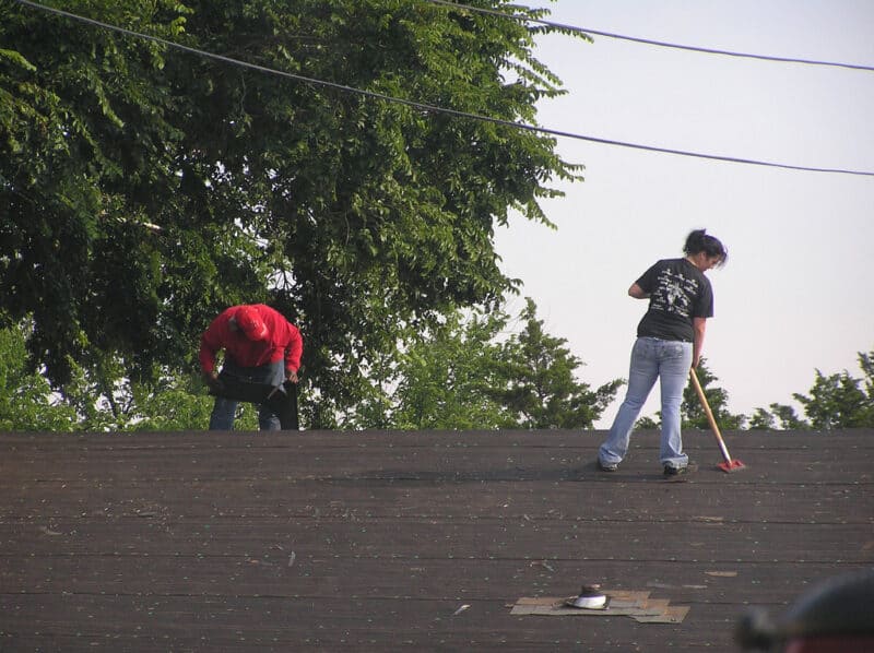 Two diverse workers installing new shingles on a roof. One worker in a red shirt cuts a shingle. A woman uses a push hoe to remove nails and debris.