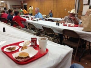 Red cafeteria tray with barbecue sandwich, beans, cookie, and drink sits on white-clothed table. Diverse diners of various ages eat and talk at long tables in community hall. Volunteers serve from kitchen area in background.