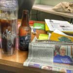 Clear coffee travel mug with iced coffee, and a local newspaper on a busy desk.