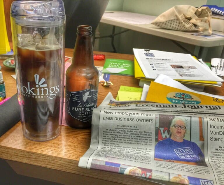 Clear coffee travel mug with iced coffee, and a local newspaper on a busy desk.