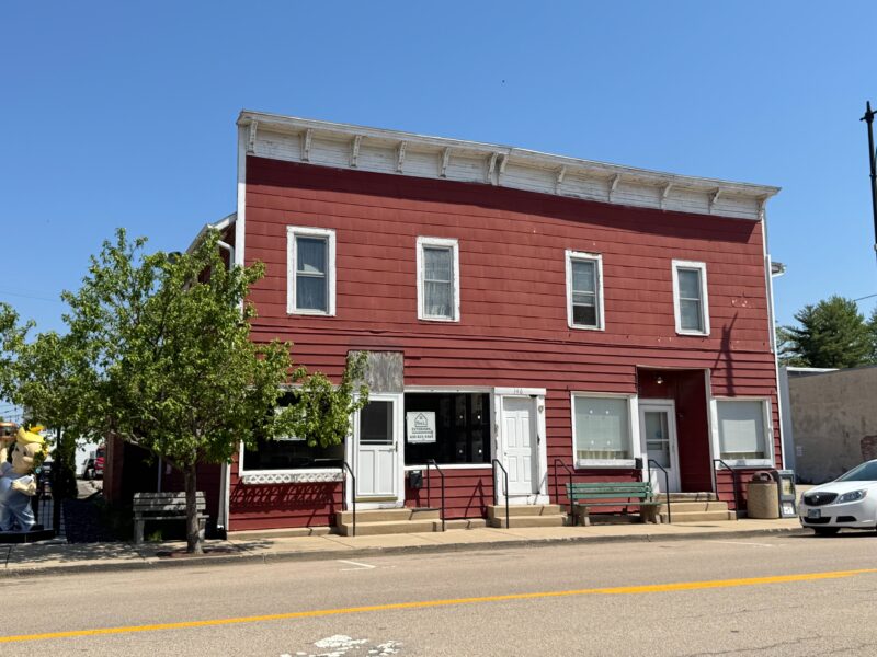 Two-story downtown building in Hinckley, Illinois with burgundy-colored siding. Three entrance doors with 2 steps in front of each and 4 picture windows. 4 small upstairs windows with old white curtains. There's a bench on the left, one in the middle and a trash can on the right. Building appears to be late 1800s or early 1900 structure. The three store fronts appear to be empty.