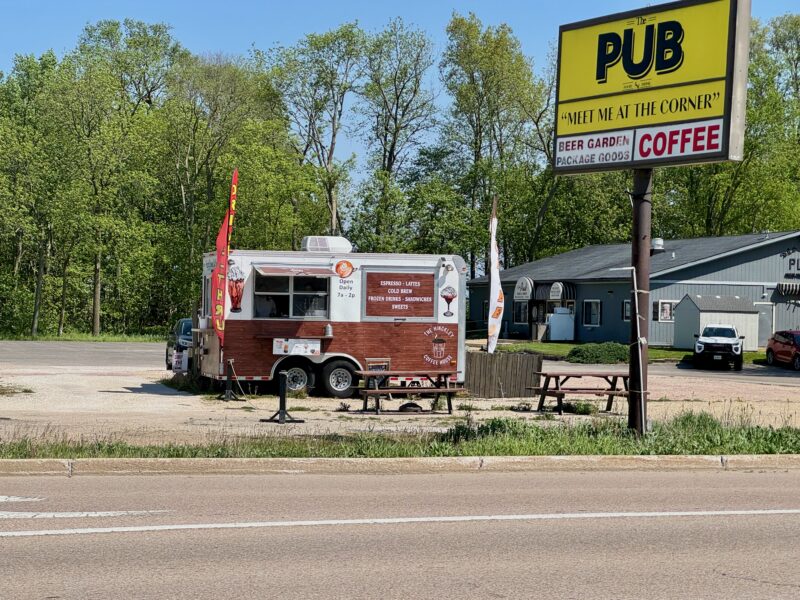 Brown and white trailer converted to a coffee shop in a parking lot. Two picnic benches in front and two flowing cloth signs on poles. Grass along curb needs trimmed. There is a sign along the roadway with The Pub in black on a yellow background and "meet at the corner" under that. At the bottom of the sign are 'beer garden' and 'coffee' in red letters on a white background. at the back of the parking lot on the right is a gray one story structure that appears to be the pub.