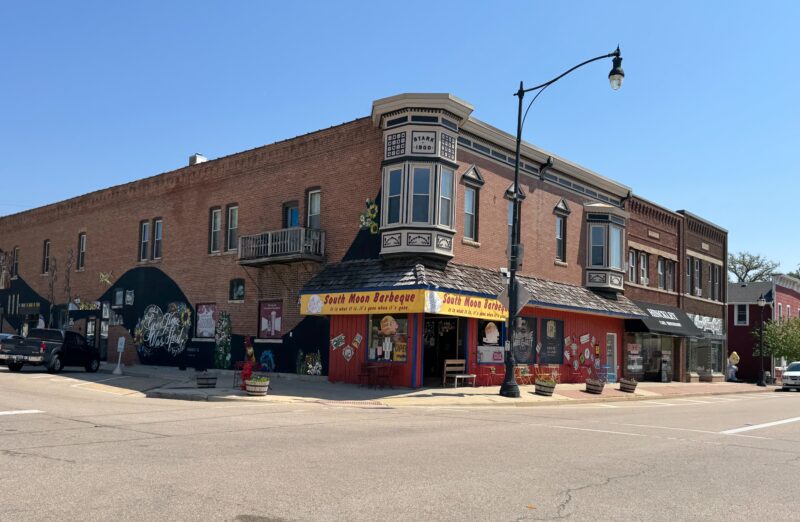 Historic two story brick corner building on a sunny day with South Moon Barbeque on the ground floor, bright red storefront and yellow awning, colorful murals and signs along the sidewalk, and neighboring small-town Main Street shops in the background in the town of Hinckley, Illinois. Second story has 2 projecting bays and over 15 small windows and could be second story housing