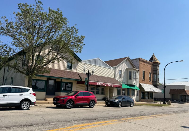 4 buildings in a row and three appear to be empty, on a street in downtown Waterman, Illinois. The full building is Sacc O Suds laundromat with several cars parked in front. The building on the end of brick with a turret and second story housing. The other three aretan 2 story frame, with room for retail on the bottom and picture windows.