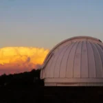 A white observatory dome against a mountainous background with sunlit storm clouds on the horizon.