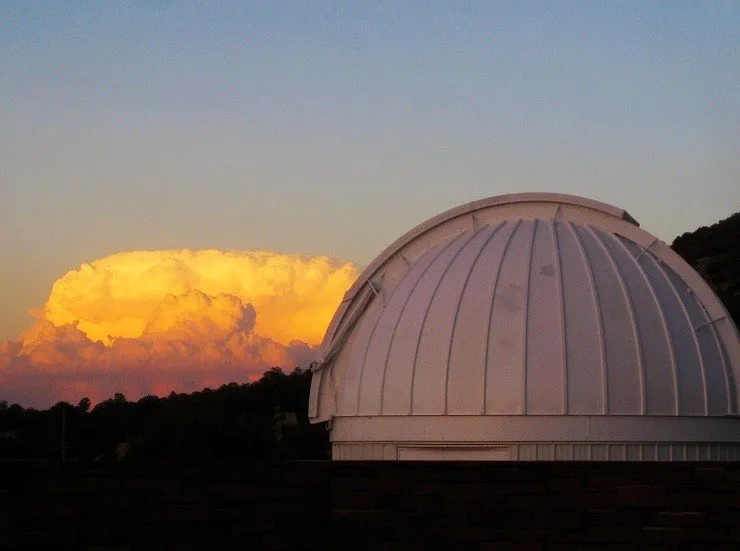 A white observatory dome against a mountainous background with sunlit storm clouds on the horizon.